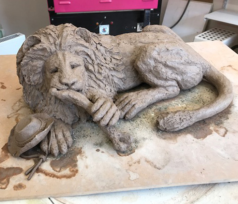 clay sculpture of a lion lying down with its paw on a bone and a hat nearby showcasing craftsmanship and detail in art design 2 features 2 textures